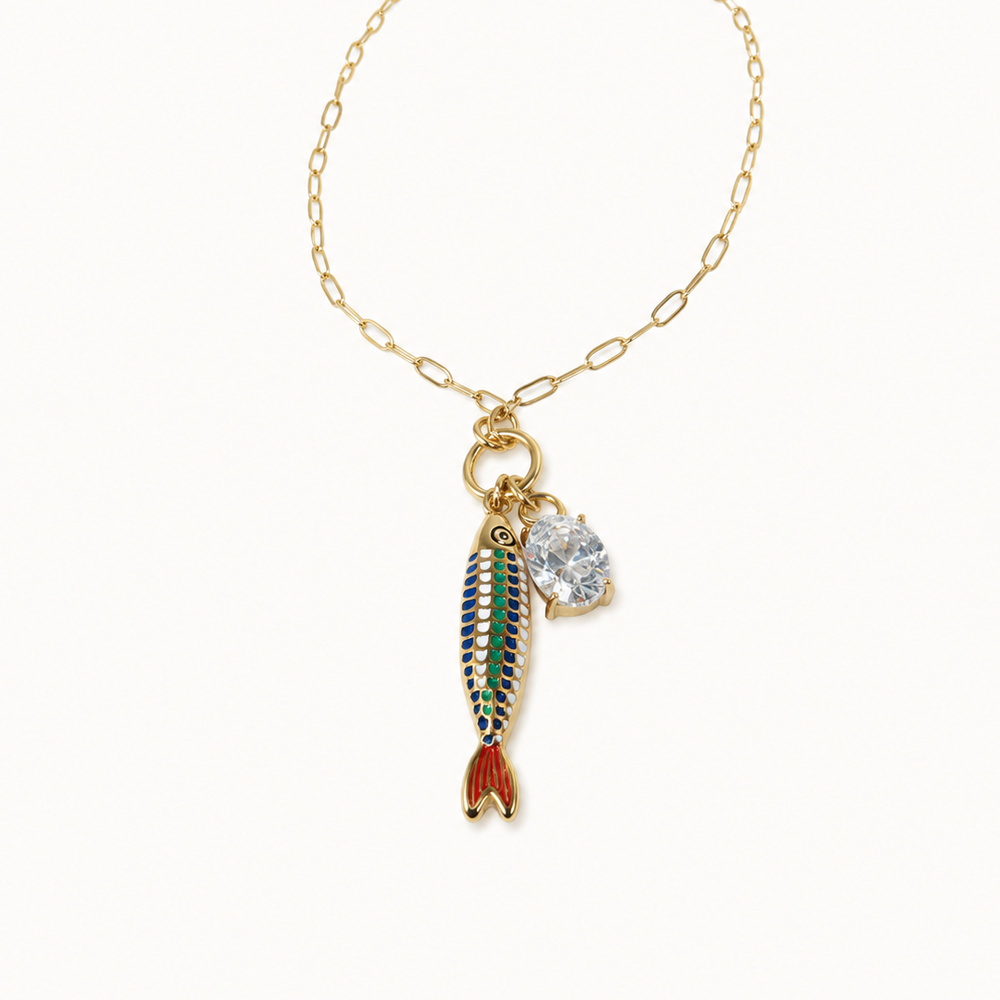 A gold chain necklace featuring a dangling fish charm with green, blue, and red enamel detailing, alongside a small clear rhinestone charm, set against a plain off-white background.