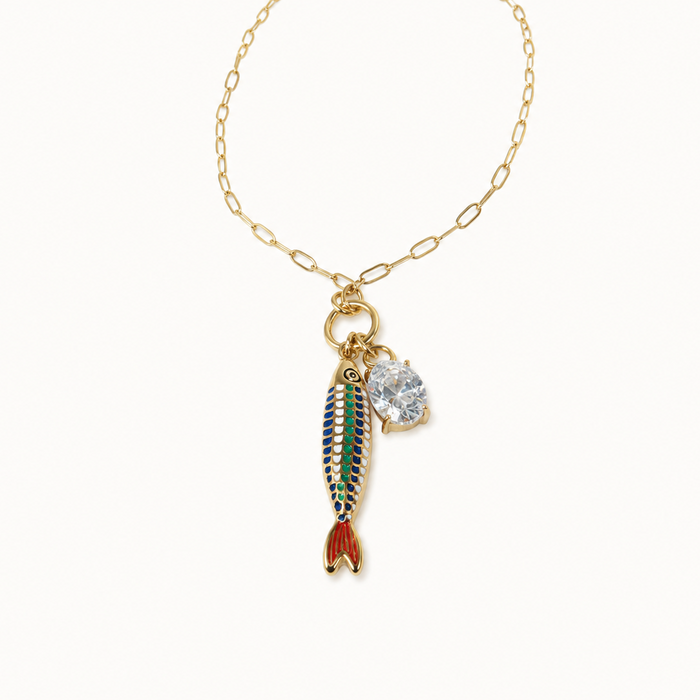 A gold chain necklace featuring a dangling fish charm with green, blue, and red enamel detailing, alongside a small clear rhinestone charm, set against a plain off-white background.