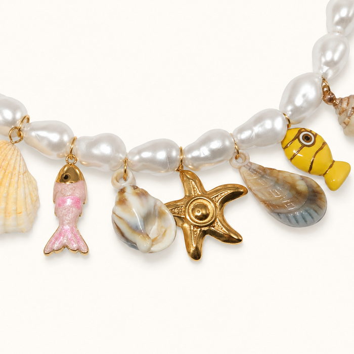A close-up of a beaded necklace with white pearl-like beads and dangling shell and fish charms, including a pink mermaid tail, a gold starfish, a yellow fish, and a blue-gray shell, on a light cream background.