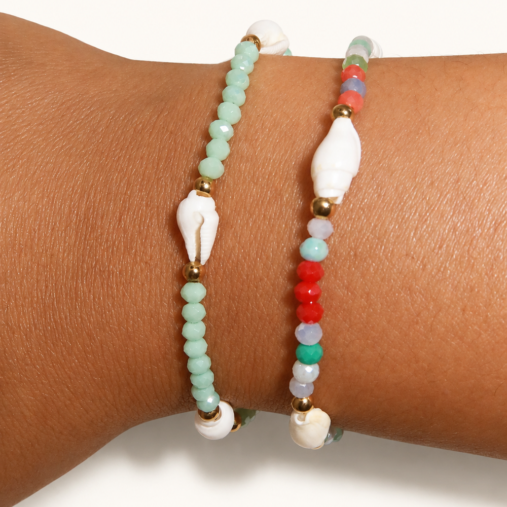 Two beaded bracelets worn on a wrist. One bracelet features light mint green beads with white seashell-shaped spacers and gold-tone beads.