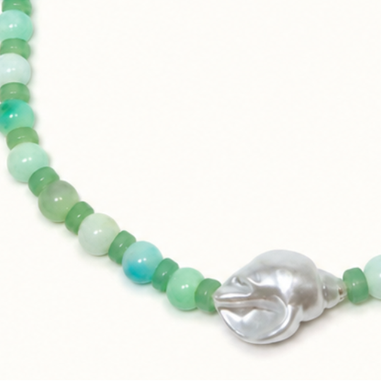 A beaded necklace featuring a mix of light green, mint, and turquoise beads, accented by a central silver-toned shell charm and a gold-toned clasp, set against a plain off-white background.