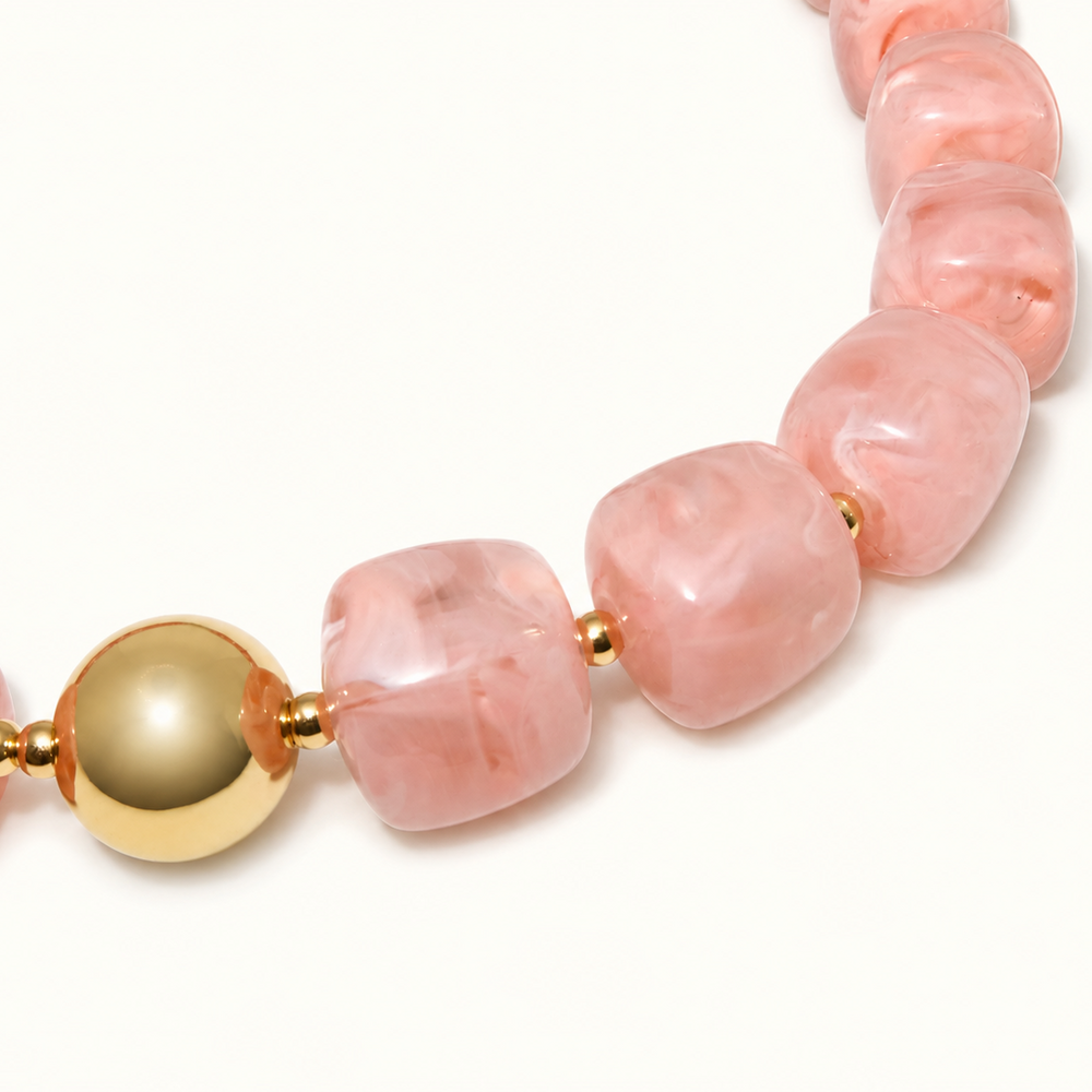 A close-up of a beaded necklace featuring chunky, polished pink stone beads and a prominent round gold bead accent, set against a clean white background.